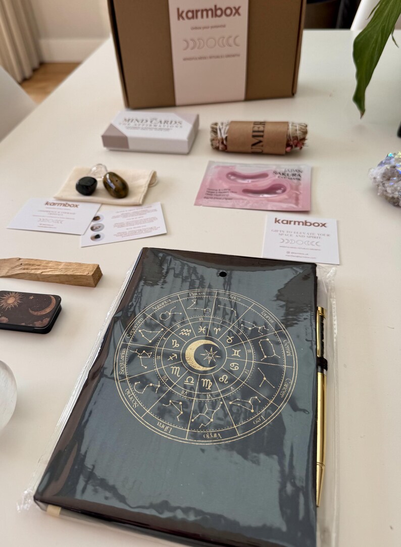 May include: A black notebook with a gold zodiac wheel design, a gold pen, a cardboard box labeled karmbox, a small box of mind cards, a bundle of dried herbs, a pink eye mask packet, two small stones in a pouch, and two informational cards.