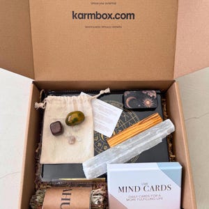 May include: A cardboard box labelled karmbox.com containing a beige drawstring pouch with three stones, a black book with a gold zodiac wheel, a selenite wand, a wooden stick, a small black tin with celestial art, and a box of LSW MIND CARDS.