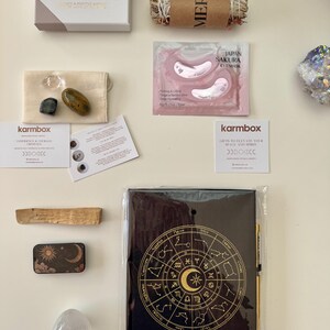 May include: A wellness gift set including LSW Mind Cards, a bundle of dried herbs labeled Merio, Japan Sakura eye masks, three small crystals in a pouch, two Karmbox cards, a palo santo stick, a sun and moon tin, a selenite egg, and a black zodiac journal.
