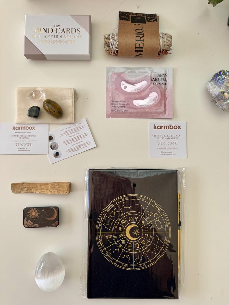 May include: A wellness gift set including LSW Mind Cards, a bundle of dried herbs labeled Merio, Japan Sakura eye masks, three small crystals in a pouch, two Karmbox cards, a palo santo stick, a sun and moon tin, a selenite egg, and a black zodiac journal.