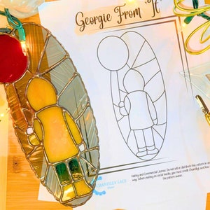 May include: A stained glass pattern of Georgie from the movie "It" holding a red balloon. The pattern is on a white sheet of paper with the words "Georgie From It" and "Digital Download" printed on it.