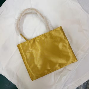 May include: A rectangular handbag in a vibrant mustard yellow satin fabric with a smooth, reflective finish. The bag features a single rounded handle that is partially covered in protective clear plastic wrapping for storage and transport.