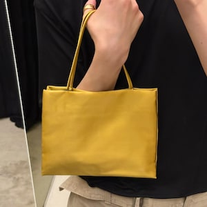 May include: A rectangular mustard yellow handbag with a smooth, satin-like finish. The bag features two thin, matching yellow shoulder straps attached to the top edge. The design is simple and structured, with clean lines and a solid, uniform color.