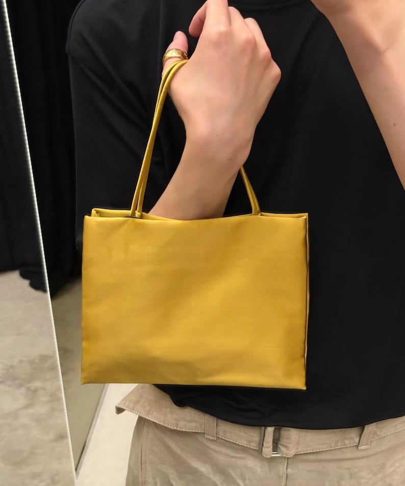 May include: A rectangular mustard yellow handbag with a smooth, satin-like finish. The bag features two thin, matching yellow shoulder straps attached to the top edge. The design is simple and structured, with clean lines and a solid, uniform color.