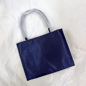 May include: A rectangular navy blue tote bag with a smooth, satin-like finish. The bag features two matching navy blue handles, each partially covered with a protective, translucent white plastic film for shipping and handling purposes.