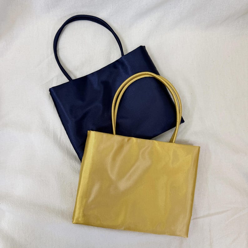 May include: Two rectangular tote bags with thin handles. One bag is a deep navy blue color and the other is a metallic gold color. Both bags feature a smooth, slightly reflective fabric finish and a simple, clean design with no external pockets.