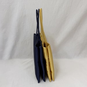 May include: Two rectangular tote bags standing upright side by side. One bag is dark navy blue and the other is a bright golden yellow. Both bags feature thin, matching fabric handles extending from the top edge, showcasing a smooth texture.
