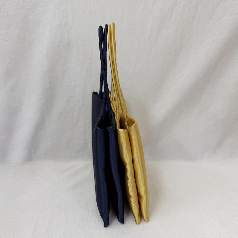 May include: Two rectangular tote bags standing upright side by side. One bag is dark navy blue and the other is a bright golden yellow. Both bags feature thin, matching fabric handles extending from the top edge, showcasing a smooth texture.