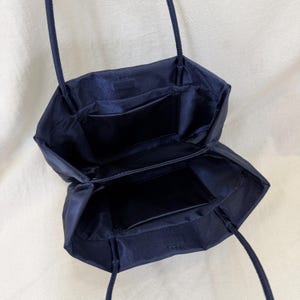 May include: A dark navy blue tote bag with a smooth, satin-like finish. The bag features an open top, two thin shoulder straps, and two internal pockets with rectangular openings, providing a clear view of the spacious, lined interior compartment.