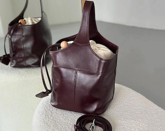 May include: A burgundy leather bucket bag with a top handle and a detachable shoulder strap. The bag has a single pocket on the front and a cream-colored lining.