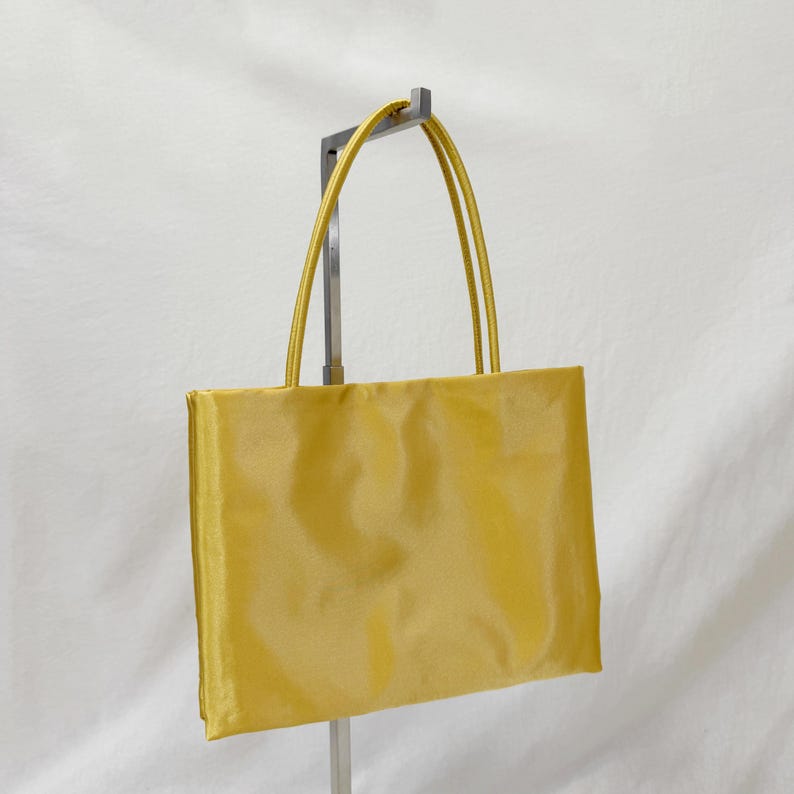 May include: A rectangular handbag in a vibrant mustard yellow color with a smooth, satin-like finish. The bag features two thin, matching yellow shoulder straps attached to the top edge, providing a simple and clean design for this accessory.