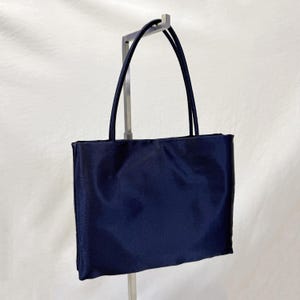 May include: A rectangular navy blue tote bag with a smooth, satin-like finish. The bag features two thin, matching navy blue shoulder straps attached to the top edge. The design is simple and structured, with a clean, solid-colored exterior surface.
