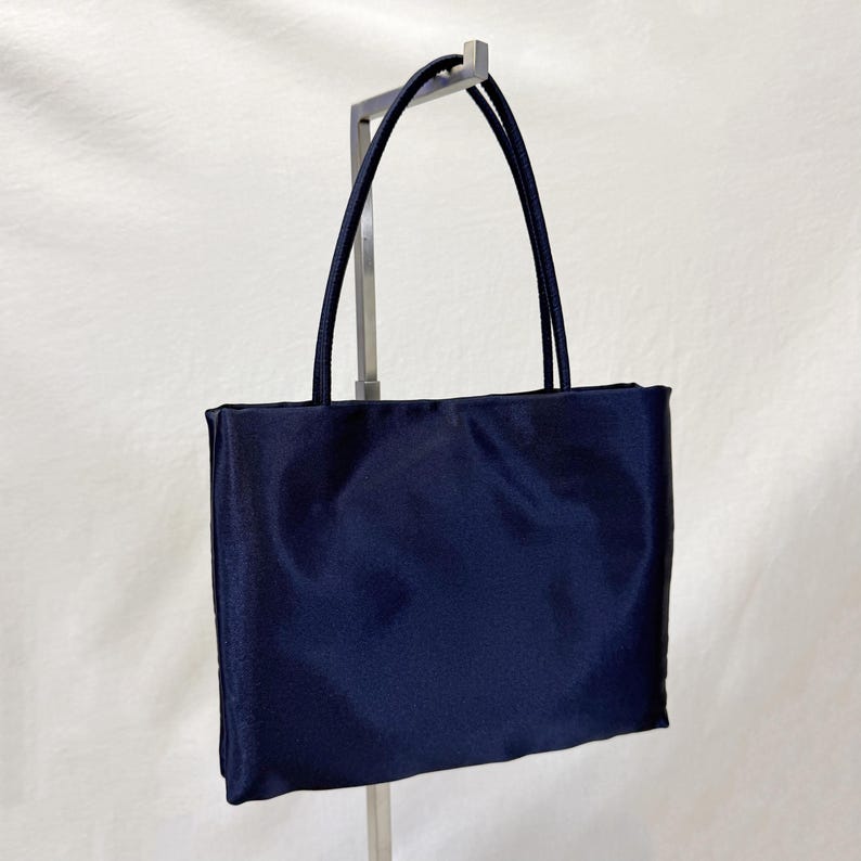 May include: A rectangular navy blue tote bag with a smooth, satin-like finish. The bag features two thin, matching navy blue shoulder straps attached to the top edge. The design is simple and structured, with a clean, solid-colored exterior surface.