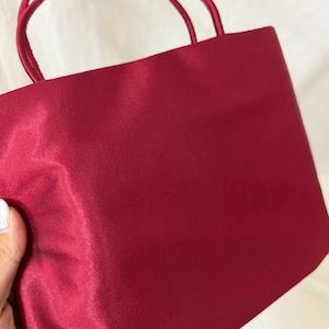 May include: A deep red handbag with a smooth, satin-like finish and two matching thin rounded handles. The bag features a rectangular body with clean, minimalist lines and a solid, uniform color throughout the exterior surface of the fabric.