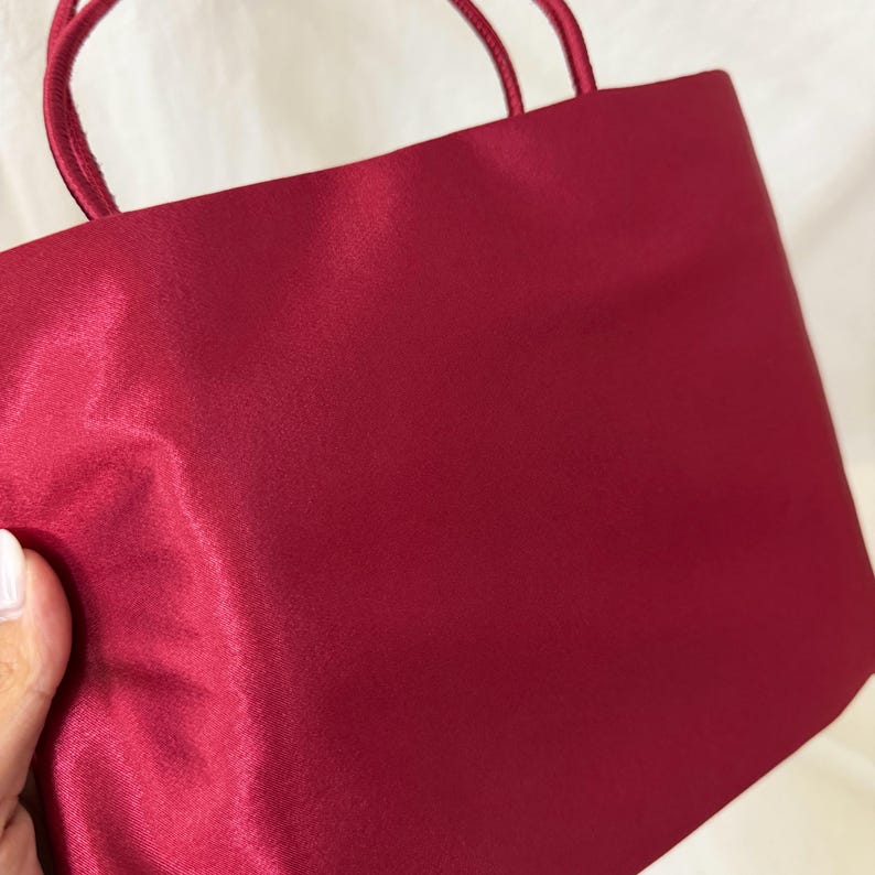 May include: A deep red handbag with a smooth, satin-like finish and two matching thin rounded handles. The bag features a rectangular body with clean, minimalist lines and a solid, uniform color throughout the exterior surface of the fabric.
