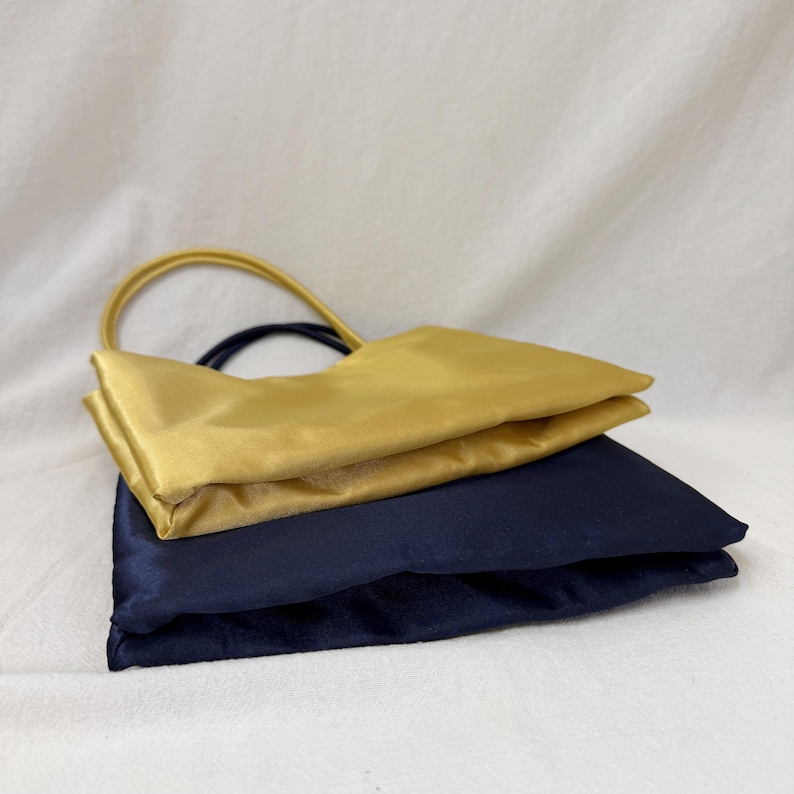 May include: A set of two rectangular satin pouches, one in a vibrant gold and the other in a deep navy blue. Each pouch features a matching circular handle attached to the top edge, with a smooth, reflective fabric finish and clean, folded seams.