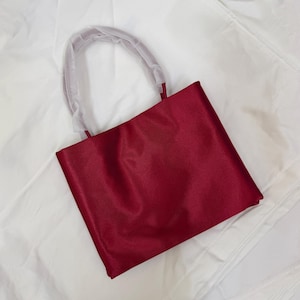 May include: A deep red satin tote bag with a rectangular silhouette and a single rounded handle. The handle is partially covered in protective white translucent plastic wrapping, maintaining a smooth and polished appearance for the fabric surface.