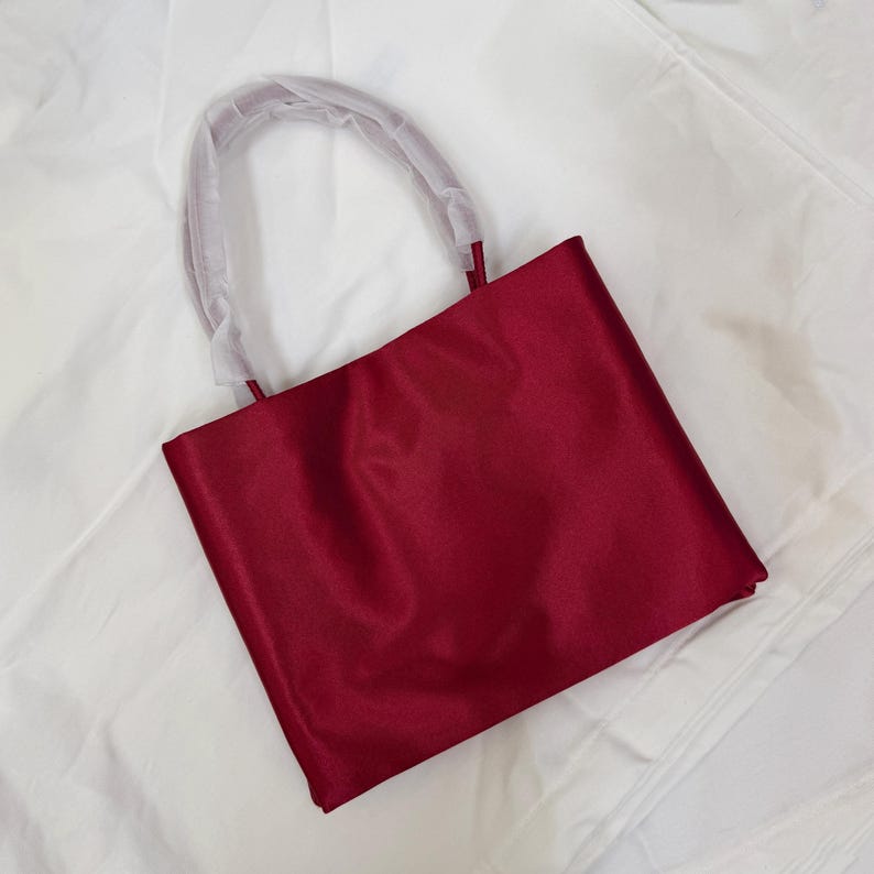 May include: A deep red satin tote bag with a rectangular silhouette and a single rounded handle. The handle is partially covered in protective white translucent plastic wrapping, maintaining a smooth and polished appearance for the fabric surface.