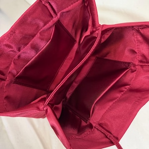 May include: A deep red satin bag interior featuring a smooth, lustrous fabric lining. The bag is open, revealing two rectangular interior pockets stitched onto the side panels, with gathered fabric detailing along the top edges and thin cord straps.