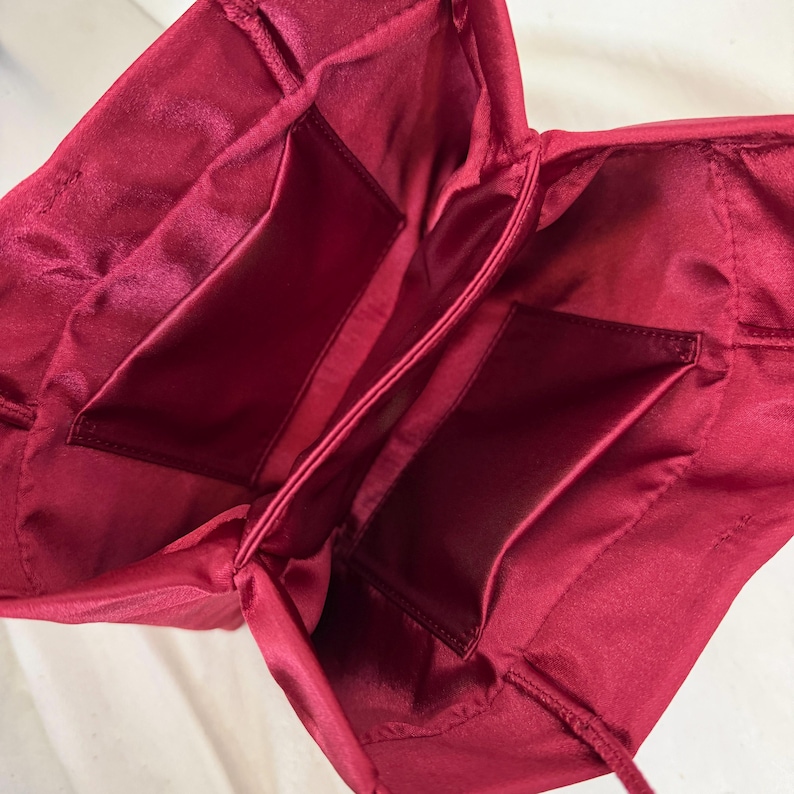 May include: A deep red satin bag interior featuring a smooth, lustrous fabric lining. The bag is open, revealing two rectangular interior pockets stitched onto the side panels, with gathered fabric detailing along the top edges and thin cord straps.