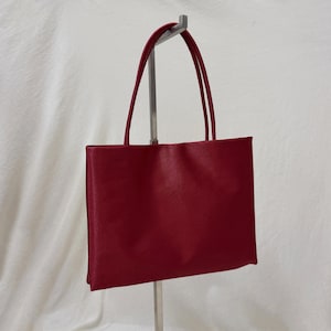 May include: A rectangular handbag in a deep red color with a smooth, satin-like finish. The bag features two thin, matching red shoulder straps attached to the top edge, providing a clean, structured silhouette for carrying personal items.
