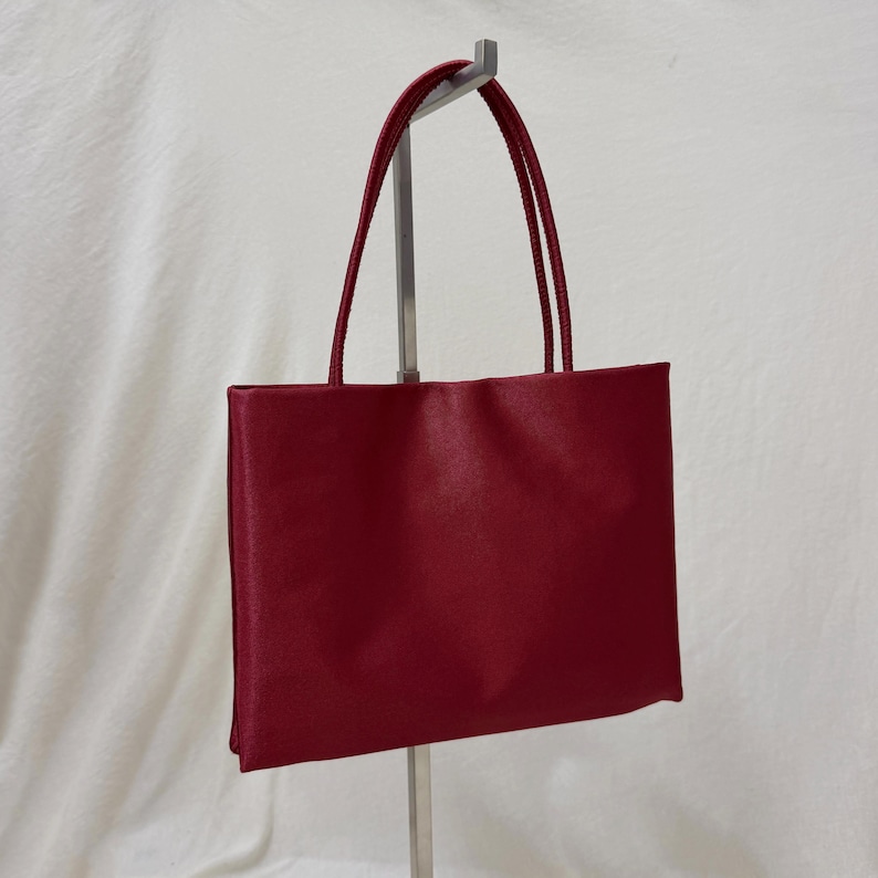 May include: A rectangular handbag in a deep red color with a smooth, satin-like finish. The bag features two thin, matching red shoulder straps attached to the top edge, providing a clean, structured silhouette for carrying personal items.