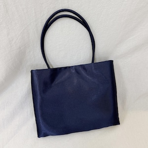 May include: A rectangular navy blue handbag with a smooth, satin-like finish. The bag features two thin, rounded top handles attached to the upper edge of the main body, providing a simple and structured design for carrying personal items.
