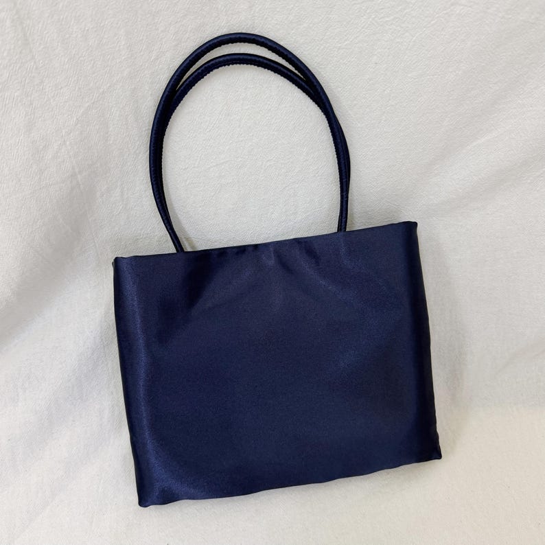 May include: A rectangular navy blue handbag with a smooth, satin-like finish. The bag features two thin, rounded top handles attached to the upper edge of the main body, providing a simple and structured design for carrying personal items.