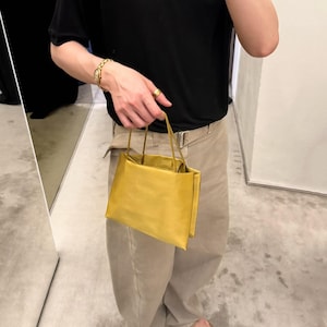 May include: A small, rectangular, mustard-yellow handbag with a smooth, matte finish. The bag features two thin, matching yellow top handles and a structured, boxy silhouette with clean, sharp edges and a simple, minimalist design aesthetic.