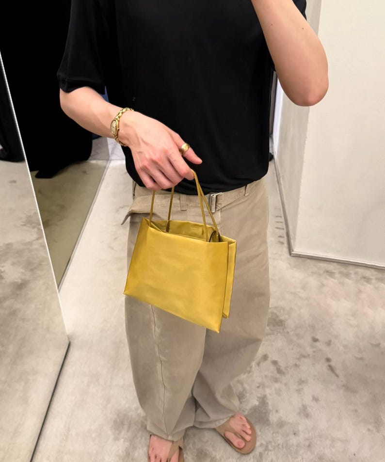 May include: A small, rectangular, mustard-yellow handbag with a smooth, matte finish. The bag features two thin, matching yellow top handles and a structured, boxy silhouette with clean, sharp edges and a simple, minimalist design aesthetic.