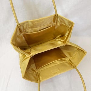 May include: A gold satin tote bag with two thin shoulder straps, featuring an open top design and a spacious interior compartment with a single rectangular slip pocket attached to the inner wall, showcasing a smooth, lustrous fabric finish.