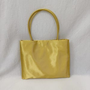 May include: A rectangular handbag in a solid, lustrous gold satin fabric. The bag features two matching gold satin top handles that are securely attached to the upper edge of the main body, creating a simple, structured, and uniform appearance.