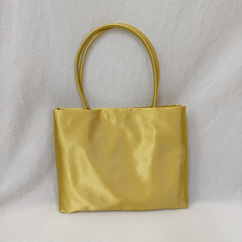 May include: A rectangular handbag in a solid, lustrous gold satin fabric. The bag features two matching gold satin top handles that are securely attached to the upper edge of the main body, creating a simple, structured, and uniform appearance.