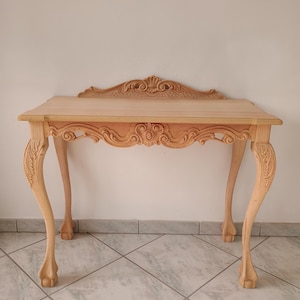May include: A light wood console table with intricate carvings on the top and legs. The table has a rectangular top and four legs that are curved and end in a claw foot design.