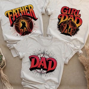 Può includere: Tre magliette bianche con grafiche. Una dice "FATHER" in rosso e giallo, un'altra dice "GIRL DAD", e la terza dice "DAD". Ogni disegno presenta un'esplosione di stelle e una silhouette.