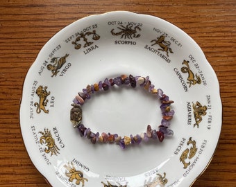 May include: A white ceramic plate with a gold rim and a zodiac wheel design. The plate features the twelve zodiac signs with their corresponding dates. A purple and brown gemstone bracelet is resting on the plate.