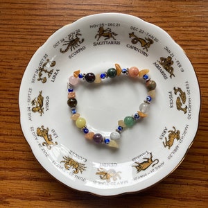 May include: A white ceramic plate with a zodiac wheel design featuring the astrological signs and dates. A colourful beaded bracelet with various stones and charms is placed on the plate.