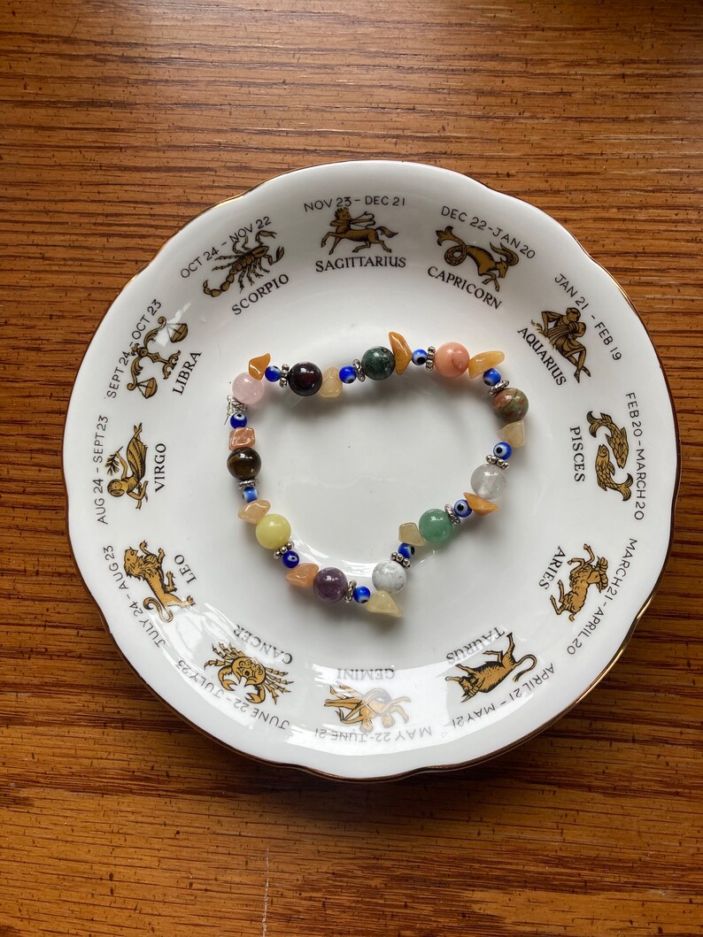 May include: A white ceramic plate with a zodiac wheel design featuring the astrological signs and dates. A colourful beaded bracelet with various stones and charms is placed on the plate.