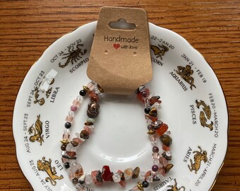 May include: Two beaded bracelets with various coloured stones, including red, brown, and clear, on a white saucer with a zodiac design. The saucer has the text "Handmade with love" on a brown tag.