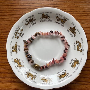 May include: A pink and brown gemstone bracelet with a gold charm, displayed on a white plate with a zodiac wheel design. The plate features the astrological signs and dates for each sign.