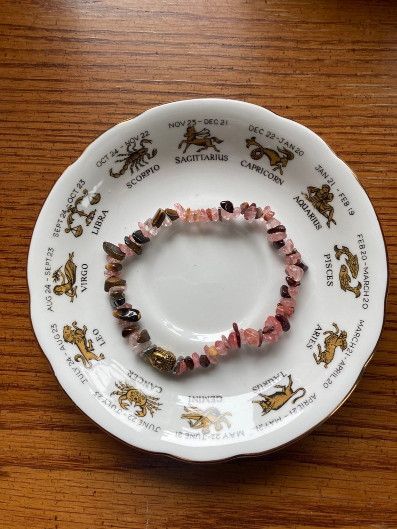 May include: A pink and brown gemstone bracelet with a gold charm, displayed on a white plate with a zodiac wheel design. The plate features the astrological signs and dates for each sign.