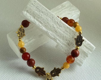 May include: A bracelet with red, orange, and yellow beads and gold hamsa hand charms. The bracelet has a small gold heart charm with an eye in the center.