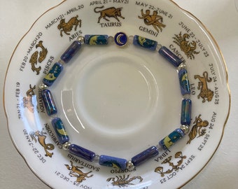 Puede incluir: Un brazalete de cuentas azules y plateadas con signos del zodíaco y fechas. El brazalete está colocado sobre un platillo blanco con borde dorado. Los signos incluyen Aries, Tauro, Géminis, Cáncer, Leo, Virgo, Libra, Escorpio, Sagitario, Capricornio, Acuario y Piscis.