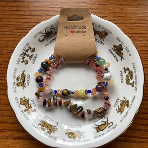 May include: Two bracelets featuring a variety of gemstone beads, including tiger's eye, rose quartz, and turquoise, displayed on a white plate with a zodiac design. The tag reads "Handmade with love".