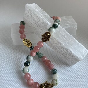 Puede incluir: Un brazalete hecho con cuentas de &aacute;gata rosa, verde y blanca y dijes de hamsa dorados.