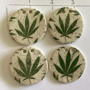 May include: Four round ceramic pendants with a green cannabis leaf design on a speckled cream background. Each pendant has two small holes at the top for stringing. The pendants are arranged in a square.