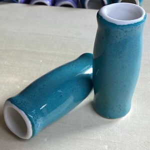 May include: Two turquoise ceramic vases with a glossy finish and white interiors. One vase is upright, the other is lying on its side. The vases have a rounded shape and a slightly flared opening. The background is a light-colored surface.
