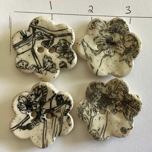 May include: Four ceramic flower-shaped buttons. Each button is off-white with a black floral design and two holes for sewing. The buttons have a speckled appearance and a slightly irregular shape.