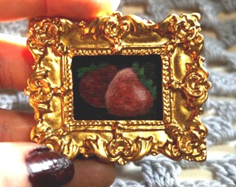 May include: A gold-toned ornate frame with a painting of two red strawberries inside. The frame has a detailed design with intricate carvings.