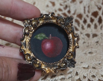 May include: A small, circular painting of a red apple with a green leaf set inside an ornate, metallic gold and dark-toned frame. The frame features intricate, raised scrollwork and decorative detailing around the circular center.